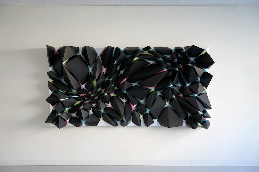 A series of black abstract shapes, each in different sizes but shaped like a black, thin and wide plinth, sticking horizontally out of a white canvas that is mounted on a wall. There is a small amount of pink and blue visible at the base of each plinth.